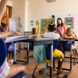 Diverse female teacher and elementary schoolchildren raising hands in class. School, learning, childhood, teaching and education, unaltered.