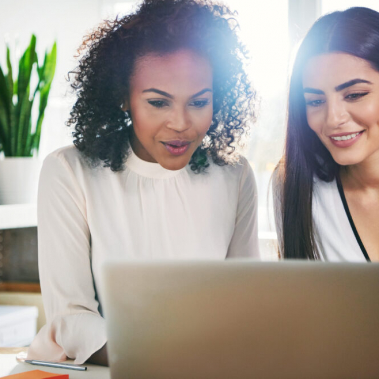 Successful dedicated young businesswomen working together on a shared laptop computer reading the screen with pleased smiles, sun flare behind