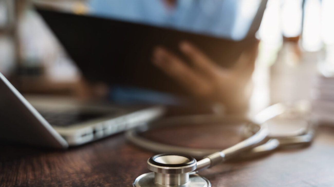Close-up of a stethoscope on a desk with a healthcare professional reviewing financial reports on a tablet, illustrating the use of financial management software in healthcare.