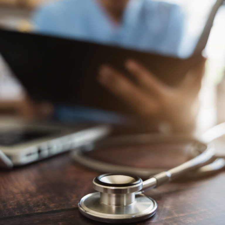 Close-up of a stethoscope on a desk with a healthcare professional reviewing financial reports on a tablet, illustrating the use of financial management software in healthcare.